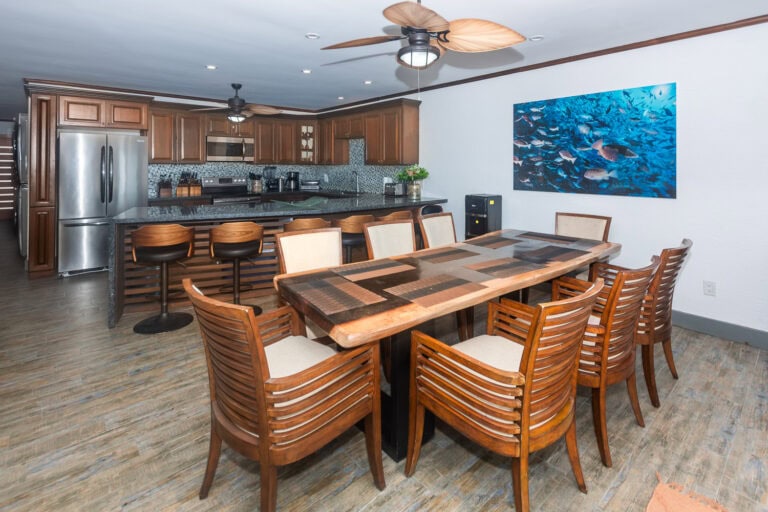Modern open-concept kitchen and dining area in a property on Roatan, Bay Islands, Honduras, featuring wooden cabinetry, stainless steel appliances, a large dining table with eight chairs, and a vibrant underwater reef photograph on the wall.