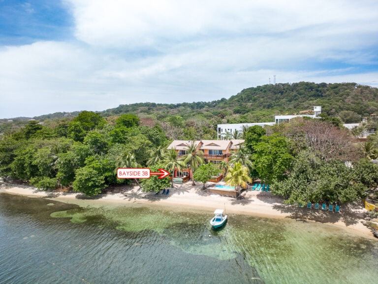 A beachfront property on Roatan in Honduras with a sign marking Bayside 3B, surrounded by lush tropical trees and clear water, with a mountain and residential buildings in the background.