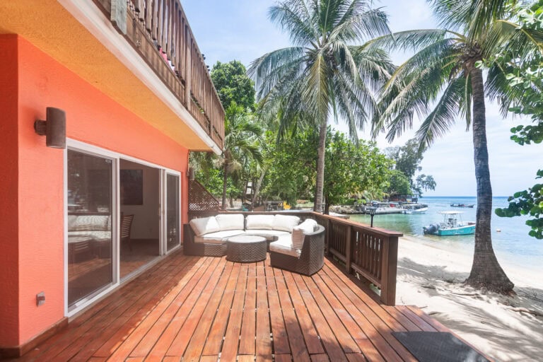 A wooden beachfront balcony with outdoor seating, including a sectional sofa and a small table, overlooking the sandy beach, palm trees, and boats docked in the clear waters of Roatan, Honduras.