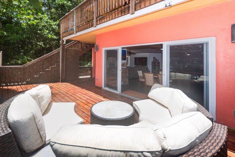 A spacious outdoor wooden deck with white cushioned seating and a round coffee table, attached to a colorful coral-colored house on Roatan in the Bay Islands of Honduras, surrounded by lush green trees.