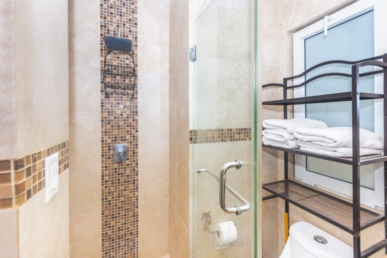 Modern bathroom in a hotel on Roatan, Bay Islands, Honduras, featuring a walk-in shower with beige tiles and a mosaic accent strip, along with a window, a towel rack holding folded white towels, and a black metal shelf