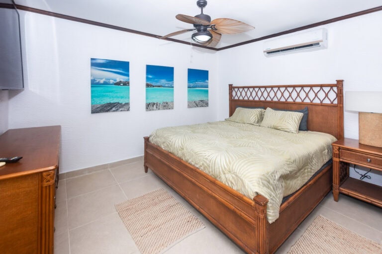 A cozy bedroom in Roatan, Honduras featuring a wooden bed with tropical-themed bedding, a ceiling fan, and three ocean-themed canvas prints on the white wall.