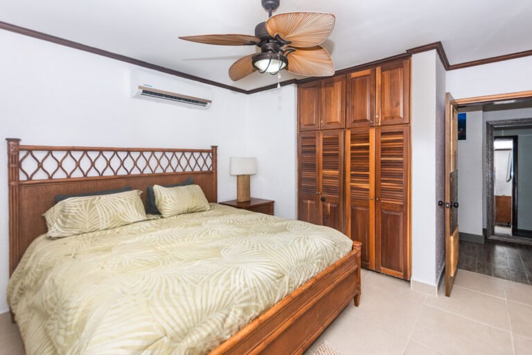 A comfortable bedroom in a Roatan hotel with a wooden bed frame, tropical-inspired bedding, a wooden wardrobe, and a ceiling fan with leaf-shaped blades, set against white walls and a Honduran coastal ambiance.