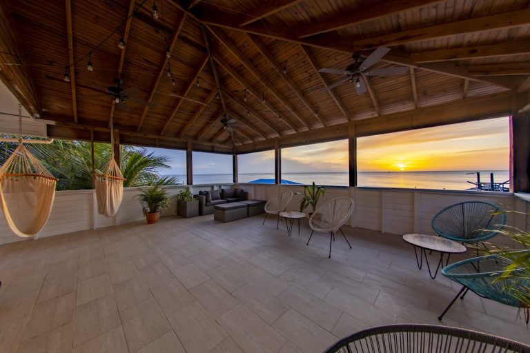 A covered outdoor lounge area on Roatan, Honduras, featuring hanging hammocks, potted plants, wicker furniture, ceiling fans, string lights, and a scenic ocean sunset view.