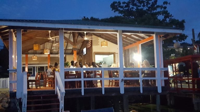 A group of people dining inside a well-lit, open-air restaurant with wooden furnishings on a raised deck in Roatan, Honduras, during the evening.