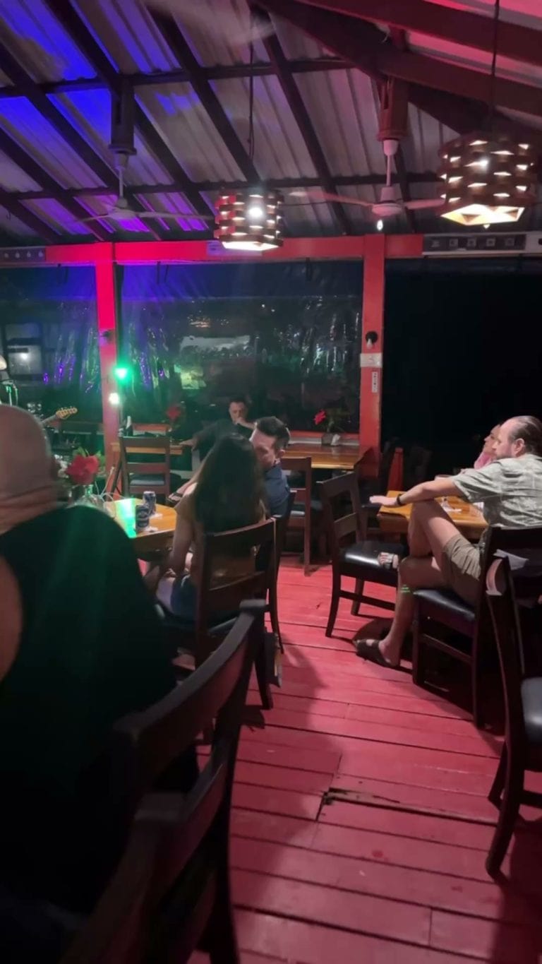 A lively indoor dining area in Roatan, Honduras, featuring wooden tables and chairs, with colorful lighting and a live music performance on stage.