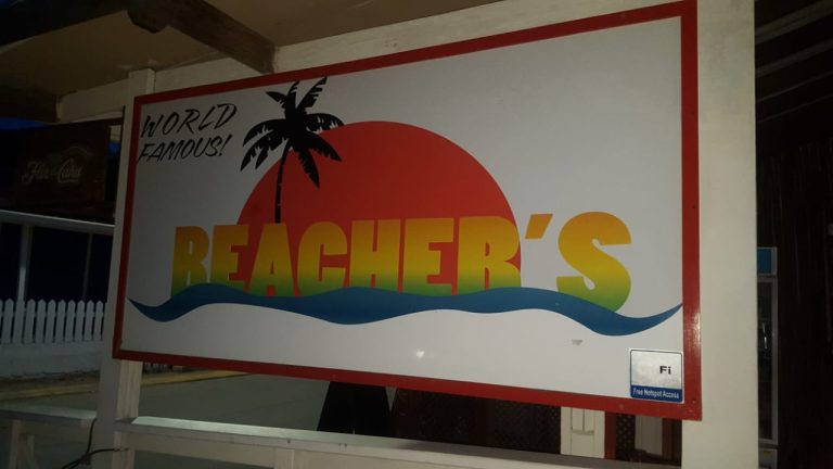 The image shows a colorful sign reading "Beacher's" with a large red sun, a black palm tree, and blue waves, indicating a beach-centric business on Roatan, Honduras.