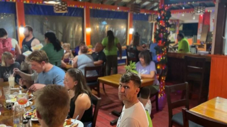 A lively restaurant in Roatan, Honduras, features diners enjoying meals at wooden tables, with colorful decor, large windows, and a Christmas tree indoors.