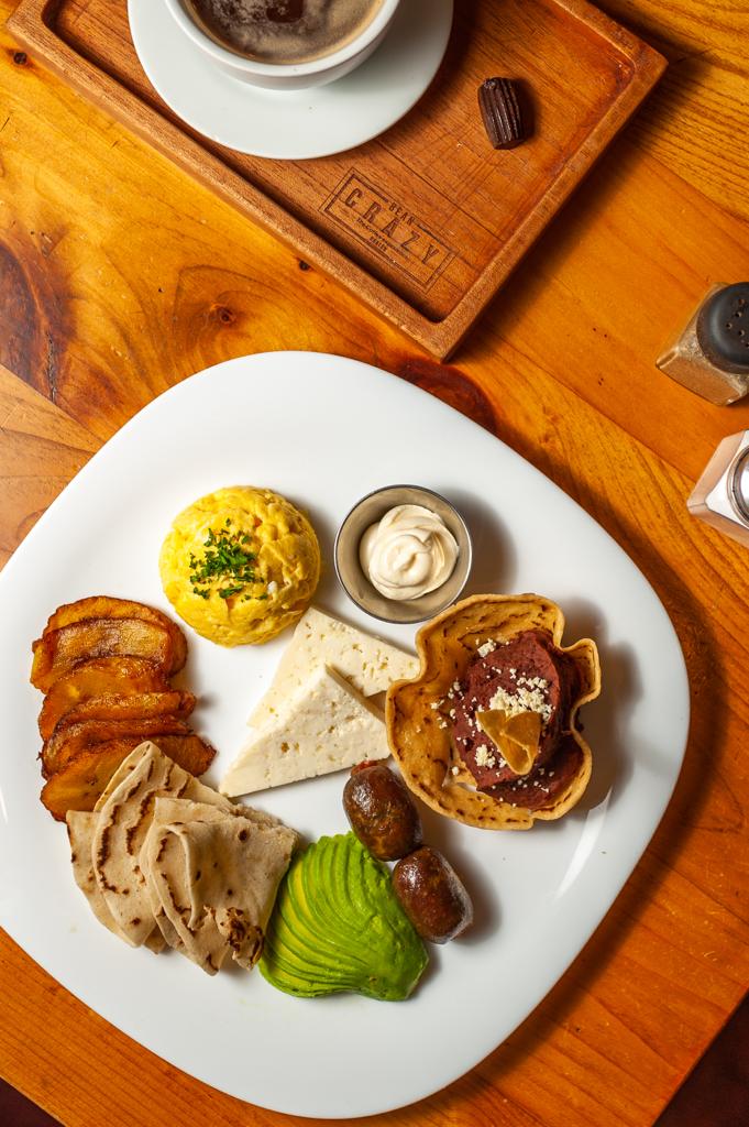 A traditional Caribbean breakfast plate from Roatan, Honduras, features scrambled eggs garnished with herbs, crispy bacon, sliced chicken or fish, mashed avocado, a small bowl of cream or sauce, grilled plantains, and a pastry topped with jam and cheese, served on a white plate on a wooden table with a cup of coffee in the background.