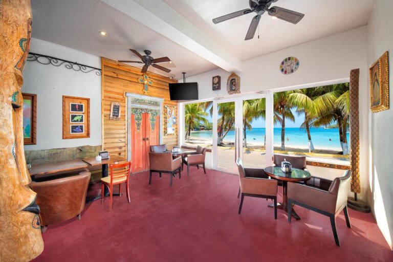 A cozy indoor seating area with brown upholstered chairs and a round glass table overlooking a beach with palm trees and clear blue water in Roatan, Honduras.