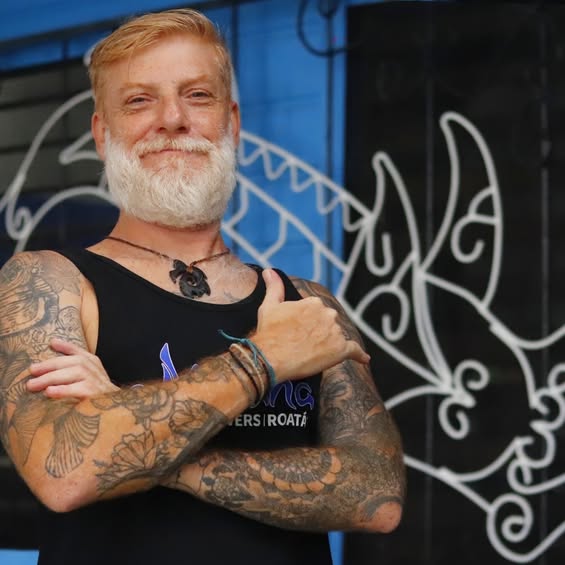 A tattooed man with a white beard and blonde hair stands confidently with crossed arms in front of a blue wall and a stylized black and white mural in Roatan, Honduras.