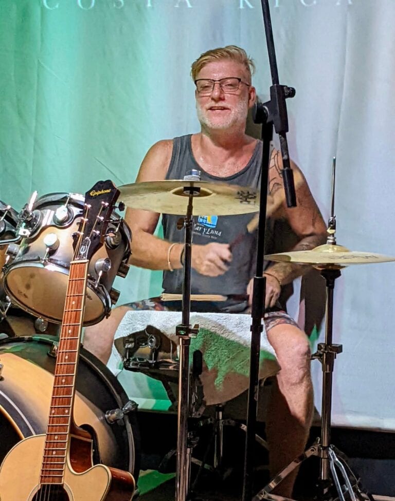 A middle-aged man with glasses and a beard plays a drum set on stage at a music event in Roatan, Honduras, with a backdrop displaying a green and white banner.