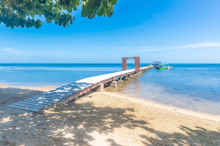 A wooden pier extends into the clear blue waters of Roatan, Honduras, with a small boat docked at the end under a shaded structure, surrounded by sandy beach and lush green foliage. The horizon shows a calm sea and a bright, sunny sky.