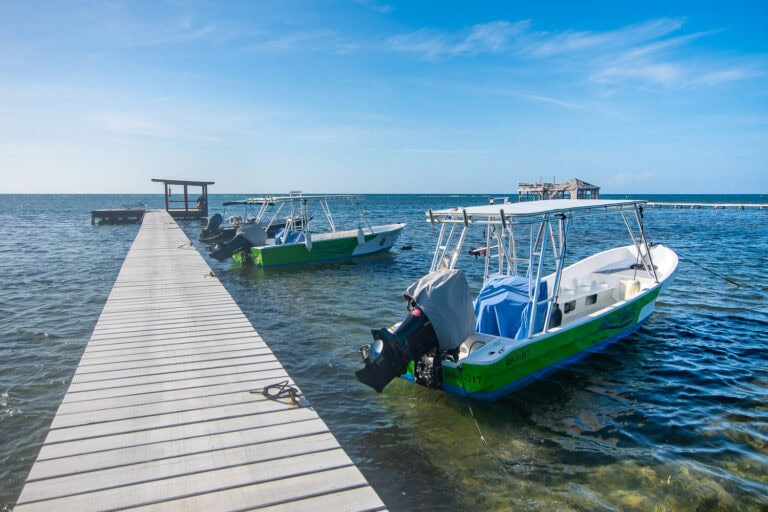 Two small green and white boats docked at a wooden pier extending into the clear blue waters of Roatan, Honduras, with a small structure in the background over the water under a bright sky.