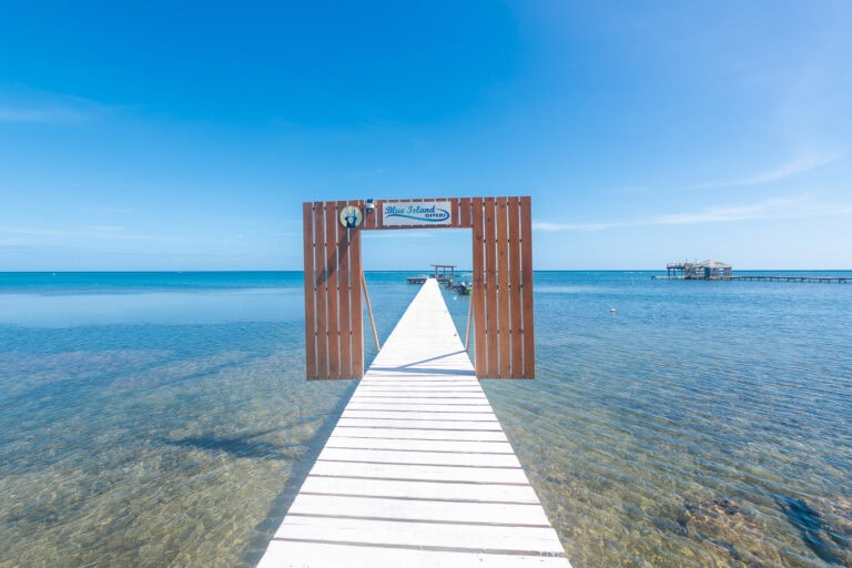 A white wooden pier extends into the clear blue waters of Roatan, Honduras, leading to a small dock with boats and a hut on stilts over the ocean.