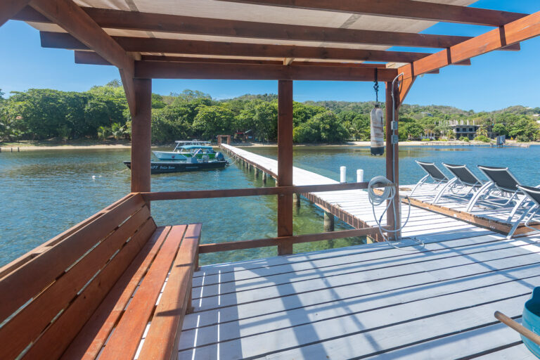 Wooden pier with lounge chairs extends into the calm waters of Roatan, Honduras, surrounded by lush green trees and a clear blue sky.