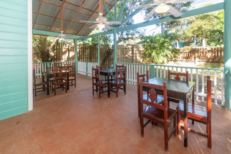 An outdoor covered seating area with wooden chairs and tables, surrounded by a white fence and lush greenery in Roatan, Honduras.