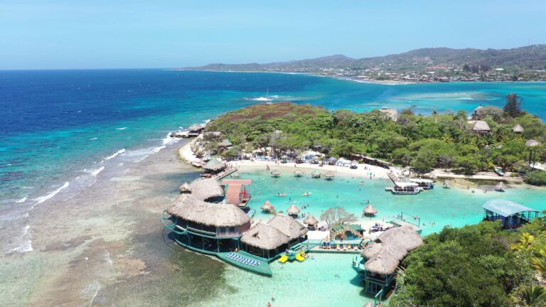 Aerial view of a beachfront resort in Roatan, Honduras, featuring turquoise waters, multiple thatched-roof structures, a swimming pool with lounge chairs, and a lush green coastline with a small town in the background.