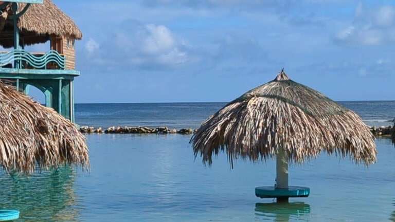 Thatched-roof umbrellas and a thatched-roof hut on stilts over the water are seen on a beach in Roatan, Honduras, with the ocean and a partly cloudy sky in the background.