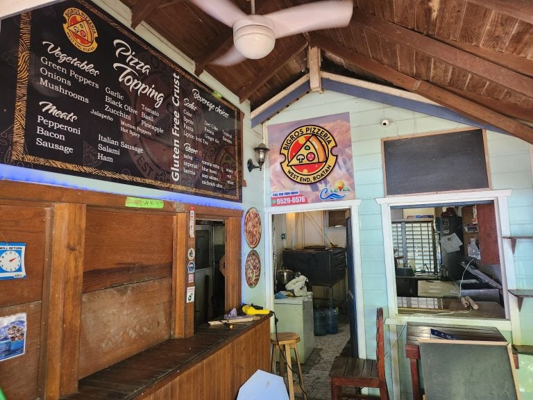 A food service counter at a Roatan, Honduras restaurant features a black menu board displaying pizza toppings, beverage options, and meat selections, with a Bigros Pizzería West End, Roatan logo and a colorful sunset mural in the background.