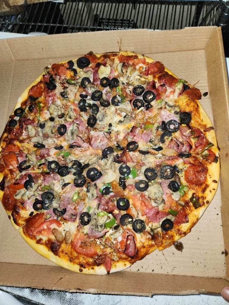 A half-eaten pizza with black olives, sliced mushrooms, green peppers, ham, and pepperoni sits inside a cardboard box on a table, with a black metal chair and some dishes visible in the background, in Roatan, Honduras.
