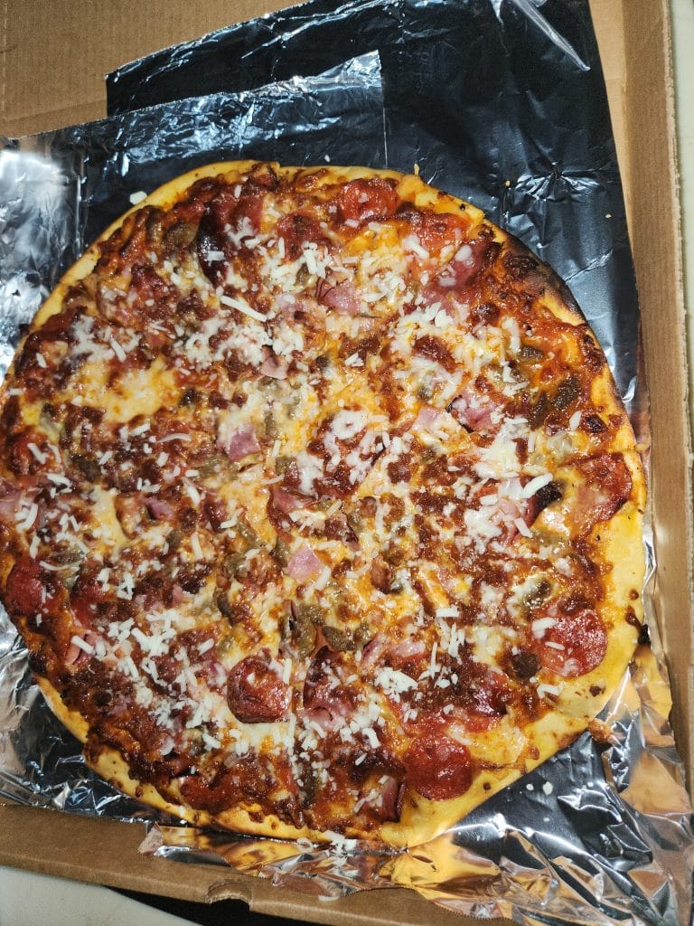 A partially sliced pizza topped with pepperoni, sausage, ham, mushrooms, and shredded cheese rests on crinkled aluminum foil inside a cardboard box on a wooden surface.