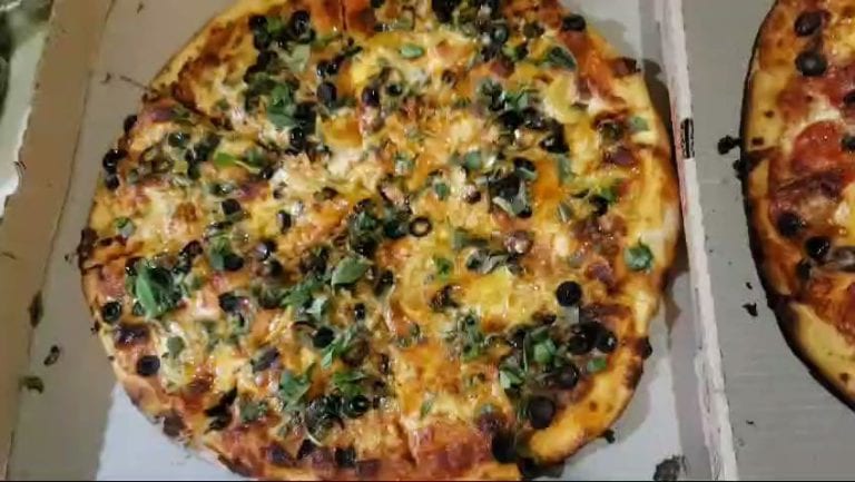A topped pizza with black olives, green herbs, and melted cheese sits in a white box, demonstrating a delicious food option available in Roatan, Honduras.