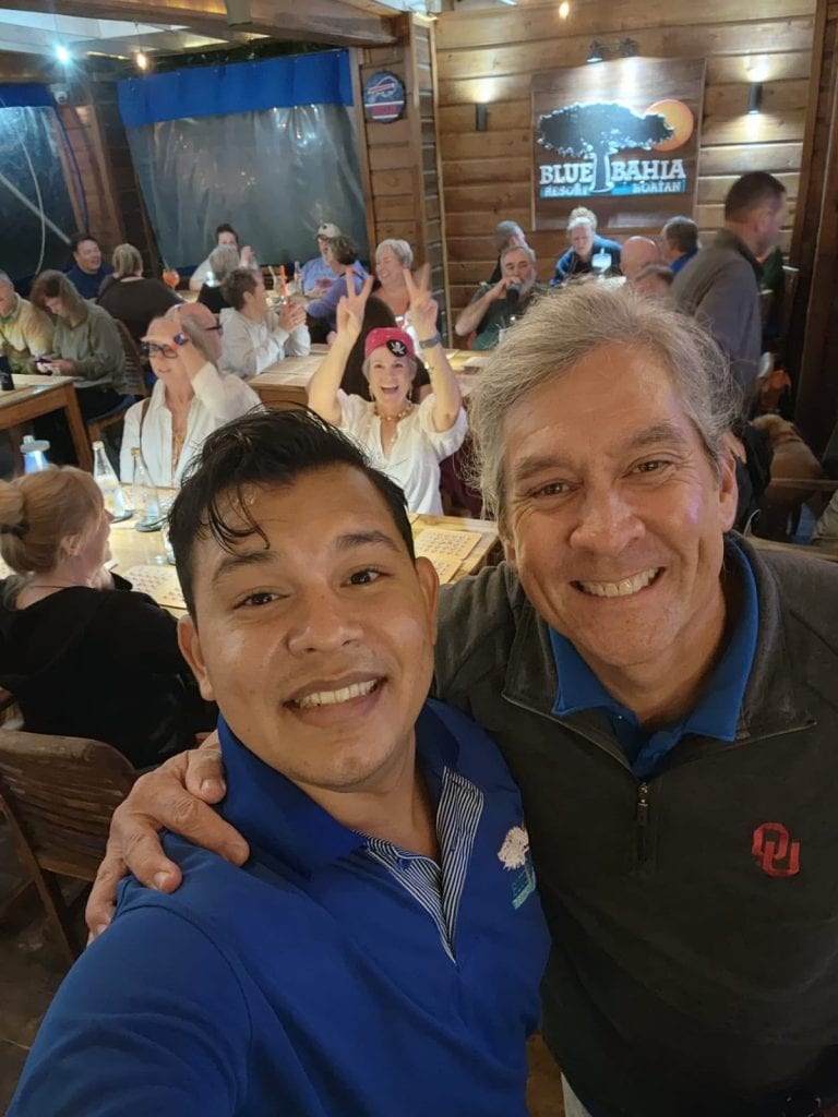 Two smiling men taking a selfie inside a lively wooden restaurant in Roatan, Honduras, with a group of people enjoying drinks and conversation in the background at Blue Bahía Resort.