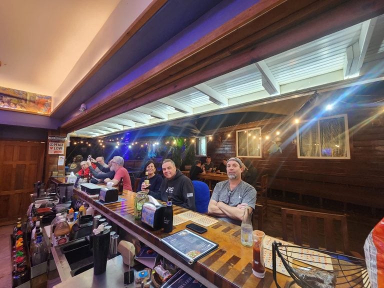 People are seated at a bar inside a wooden restaurant in Roatan, Honduras, with string lights and an open-air upper section visible.