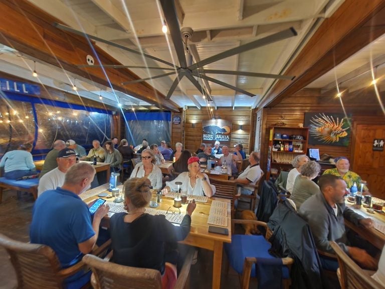 A diverse group of people dining inside a wooden-themed restaurant on Roatan, Honduras, with tropical decor, a ceiling fan, and a "Blue Bahia" sign on the wall.