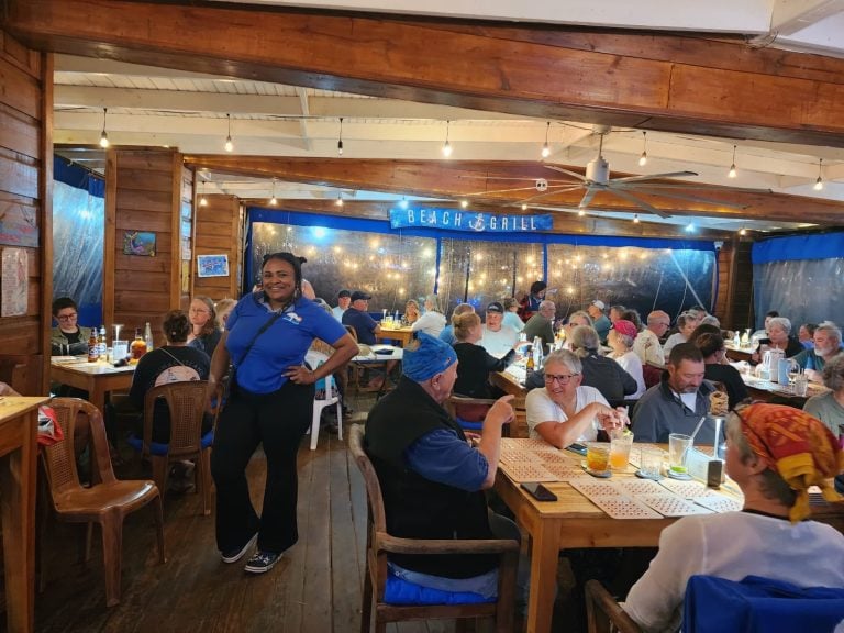 A lively indoor restaurant in Roatan, Honduras, features wooden walls, warm lighting, and a diverse crowd of diners enjoying their meals at long wooden tables with drinks, while a smiling staff member in a blue uniform stands in the foreground, with a "Beach & Grill" sign visible on the back wall.