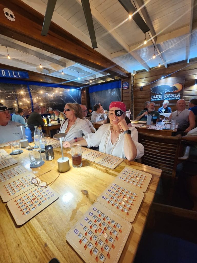 A group of people enjoying a lively evening at a wooden indoor restaurant in Roatan, Honduras, with bingo cards and drinks on the table, and a woman wearing a pirate eye patch smiling in the foreground.