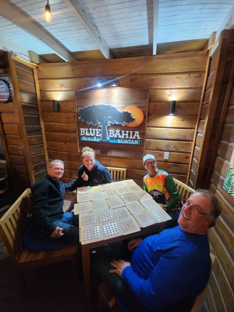 Four people enjoy a game of bingo at a wooden table inside a rustic restaurant or bar with a "Blue Bahia Resort Roatan" sign on the wooden-paneled wall, surrounded by warm lighting in Roatan, Honduras.