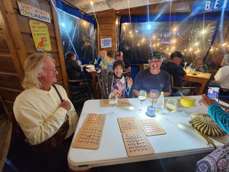 A group of people enjoying a night out at a restaurant in Roatan, Honduras, with bingo cards on the table, colorful lighting, and a cozy wooden interior featuring decorative signs and large windows revealing a nighttime scene.