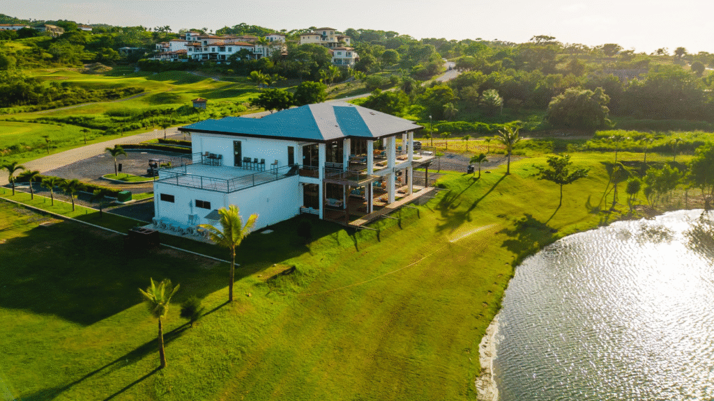 A modern three-story building with outdoor seating balconies is situated on a lush green lakeside property in Roatan, Honduras, surrounded by palm trees and overlooking a small body of water.