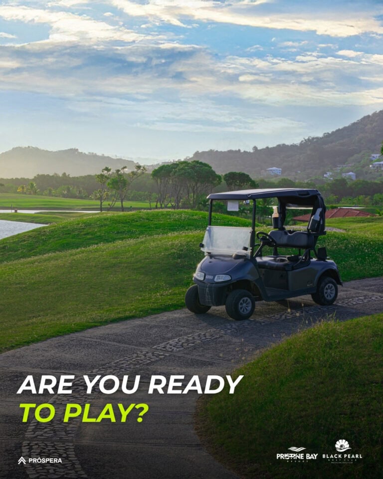 A golf cart parked on a paved path overlooks a lush green golf course with trees, water features, and rolling hills against a cloudy sky in Roatan, Honduras.