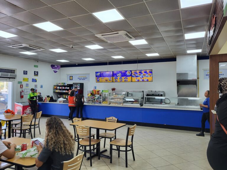 Inside a fast-food restaurant on Roatan, Honduras, customers are ordering at the counter, with digital menu boards displaying food options, while seating areas with tables and chairs are occupied by people, and staff members are assisting customers in a casual, brightly lit setting.