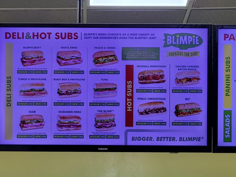 A digital menu board displaying various sub sandwiches at a saloon in Roatan, Honduras, with colorful images, prices, and ingredients listed.