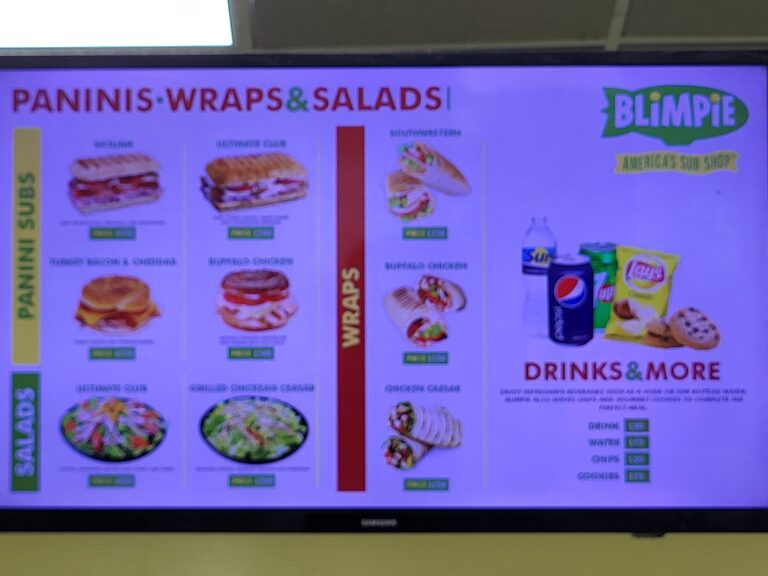 A digital screen displays a menu from a Roatan, Honduras sandwich and salad shop called Blimpie, featuring images and prices of various sandwiches, salads, and drinks.