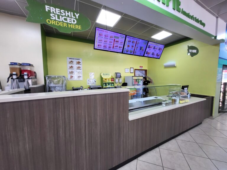 A fast-food counter inside a business on Roatan, Honduras, features digital menu screens displaying sandwiches, salads, and snacks, with signs indicating where to order and pay.