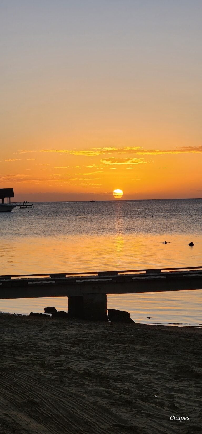 A sunset over the calm waters of Roatan, Honduras, with a wooden dock extending into the ocean and boats visible in the distance, creating a tranquil tropical scene.
