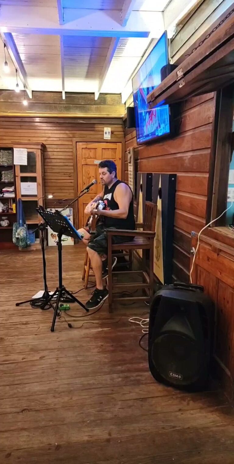 A man plays guitar and sings into a microphone inside a rustic wooden restaurant or bar on Roatan, Honduras, with a large television mounted on the wooden wall behind him.