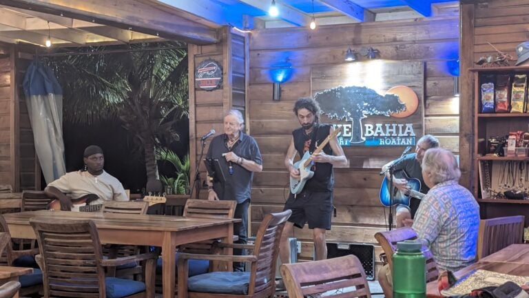 A live music performance featuring three guitarists and a singer takes place inside a wooden-walled restaurant in Roatan, Honduras, with an audience seated at tables and a tropical backdrop visible through an open window.