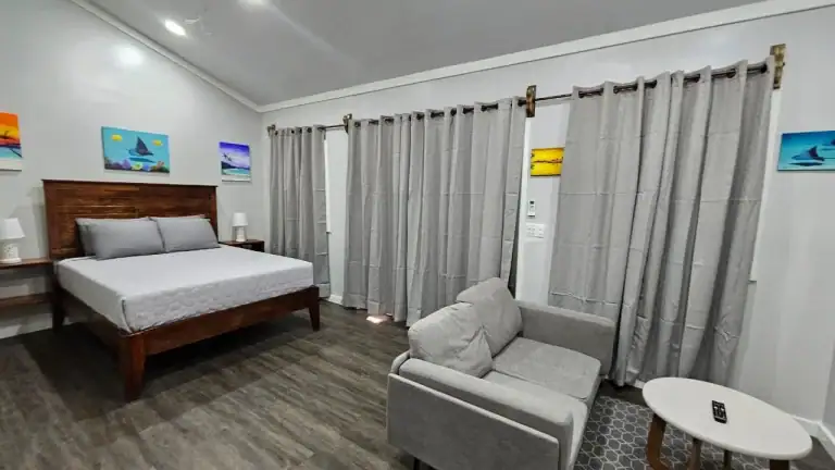 A cozy bedroom in Roatan, Honduras features a wooden bed with white bedding, artwork of sailboats and sunsets on the walls, a small gray sofa and a white oval coffee table with a remote control, and beige curtains covering the windows.