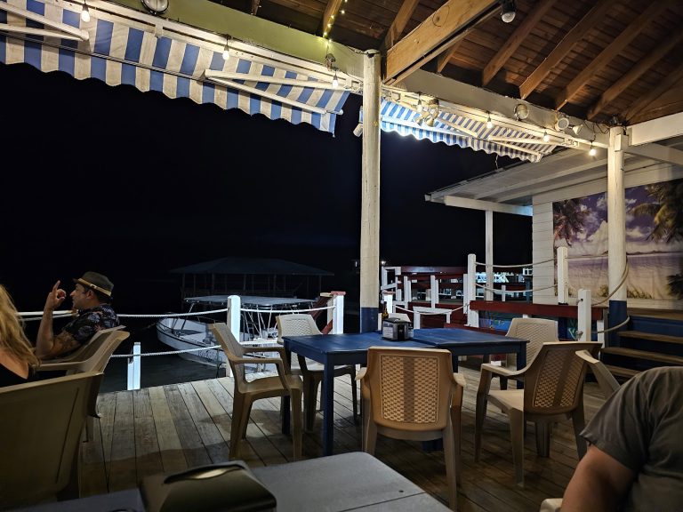 An open-air restaurant in Roatan, Honduras, features wooden decking, plastic chairs, striped blue and white awnings, decorative string lights, and a mural of a tropical beach scene on the wall.