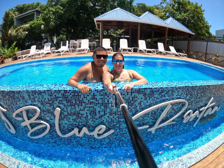A smiling man and woman are taking a selfie in a blue-tiled swimming pool at a resort in Roatan, Honduras, with lounge chairs, a covered deck area, and lush green trees in the background.