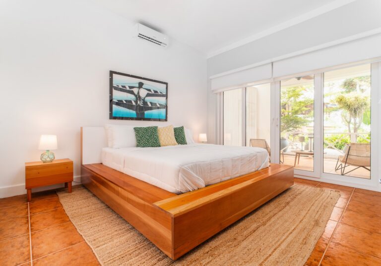 A bedroom in a Roatan hotel or vacation rental features a wooden platform bed with white bedding and decorative green and yellow pillows, with large glass doors opening to a balcony overlooking lush greenery in Honduras.