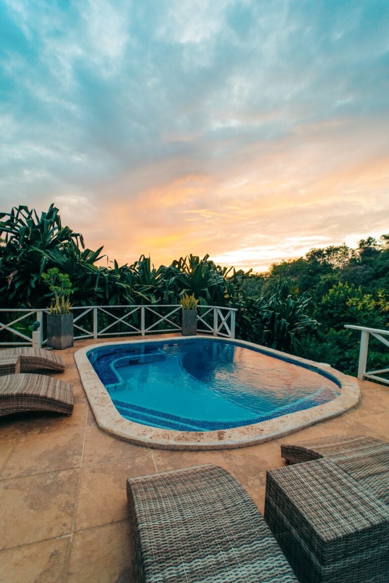 A private swimming pool surrounded by wicker lounge chairs and lush green plants overlooks a scenic sunset sky over Roatan, Honduras.