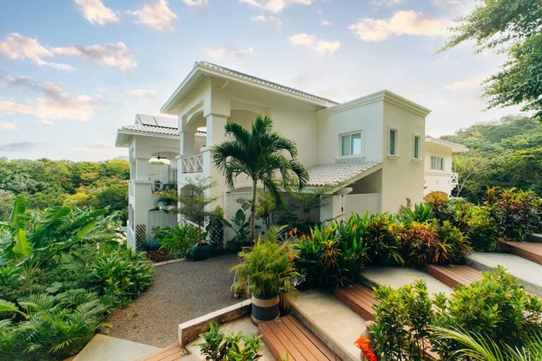 A white multi-story building with balconies and a tiled roof is surrounded by lush tropical plants and trees in Roatan, Honduras, with a clear sky and scattered clouds overhead.