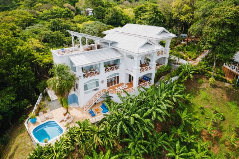 A white multi-story villa with a rooftop terrace, swimming pool, and lush tropical greenery surrounds the property on Roatan, Honduras.